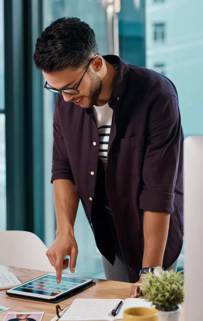 A man with glasses interacts with a tablet on a desk, surrounded by notebooks, a pen, a mug, and a small plant near a large window.