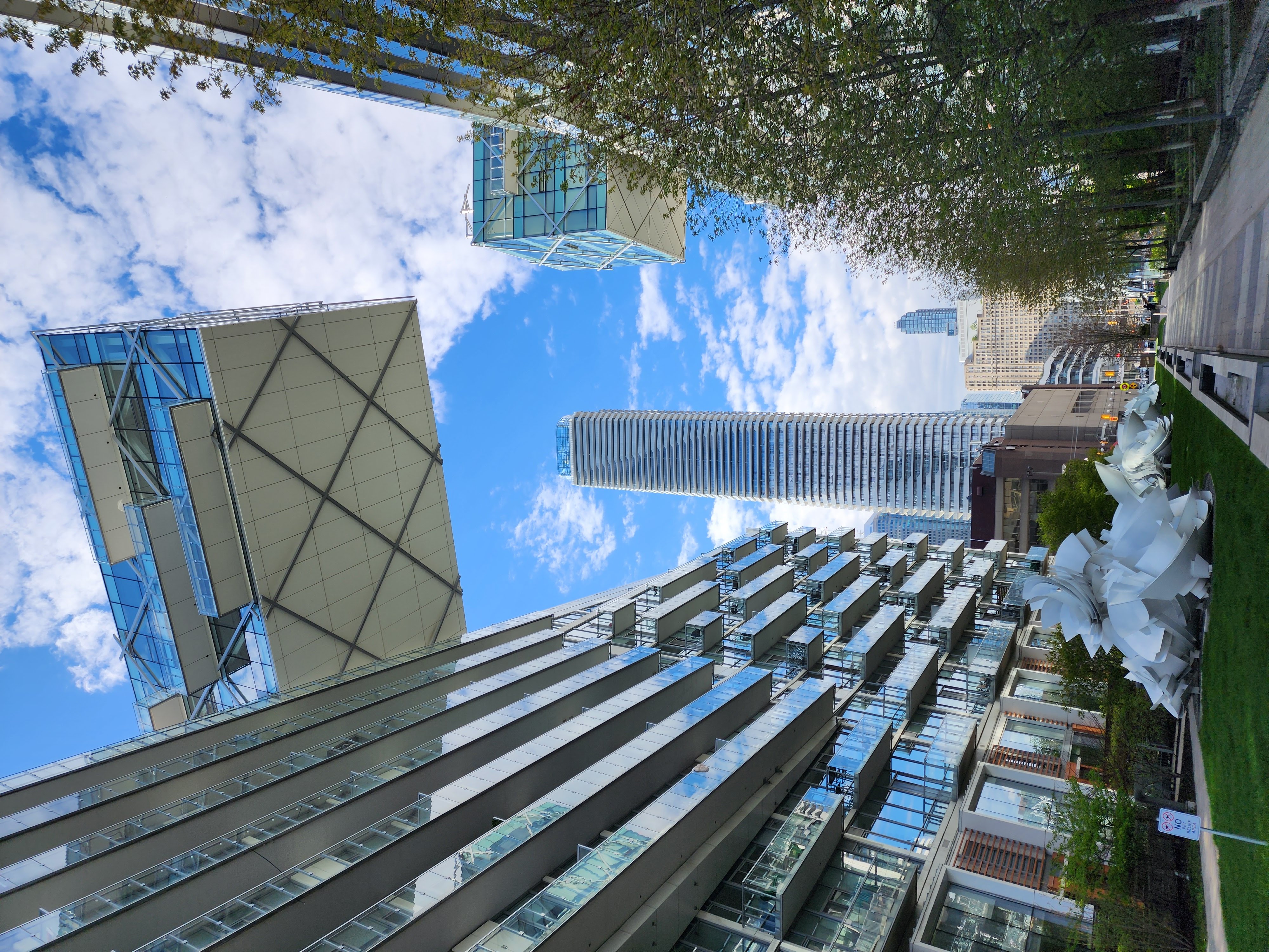 Looking up at modern glass condo towers in downtown Toronto, with a white sculpture on a lawn beside a tree-lined walkway