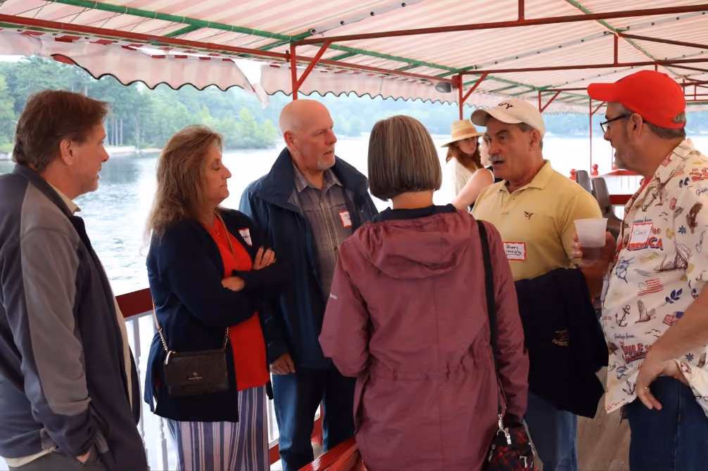 Group of six adults talking on a covered boat deck by a lake with trees in the background.