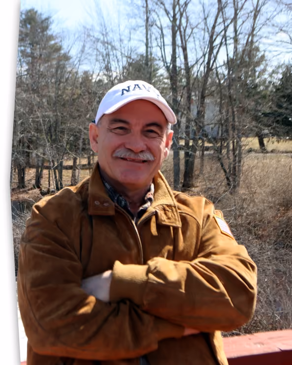 Smiling man wearing a white NAVY cap and brown jacket standing outdoors with folded arms.