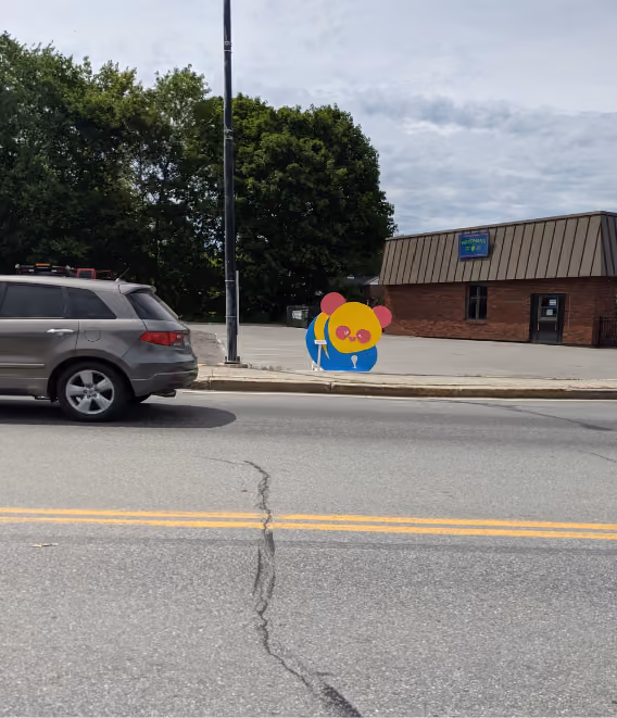 A gray SUV parked by a sidewalk with a colorful bear-shaped sign near a brick building and trees.