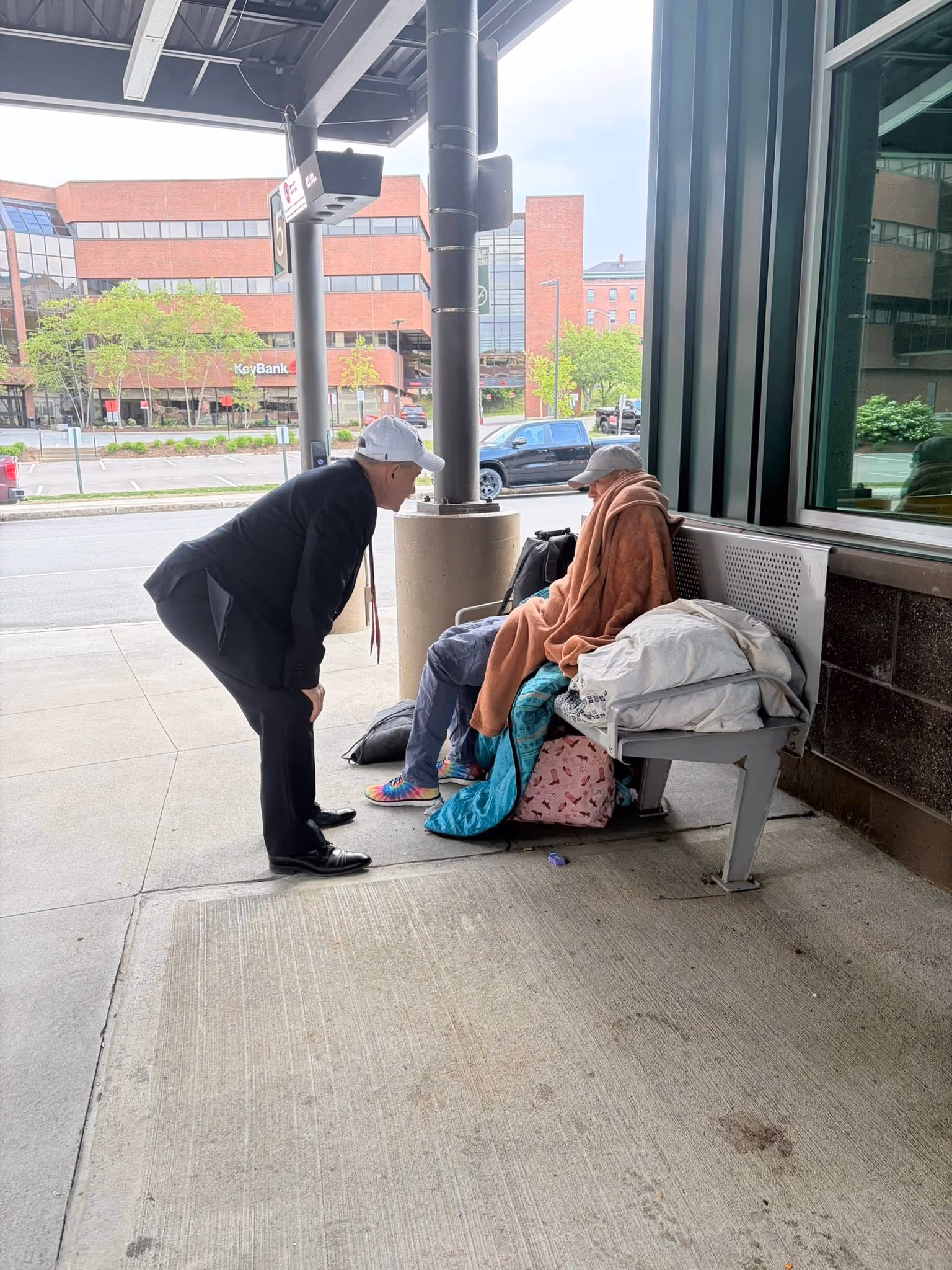 Man in a suit bends down to talk to a person wrapped in blankets sitting on a bench at a bus stop.