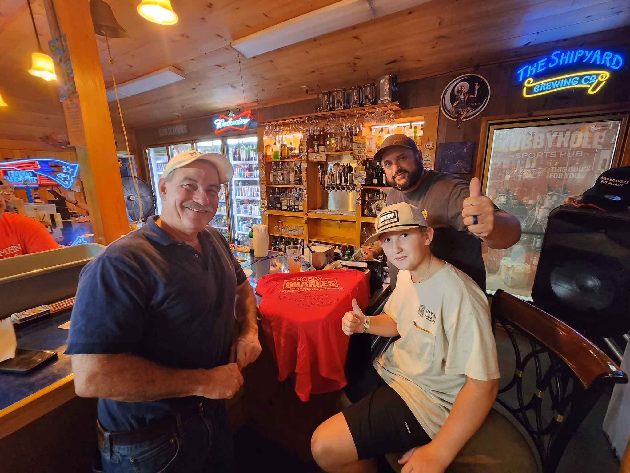 Three people inside a wood-paneled bar posing with thumbs up around a table with a red shirt.