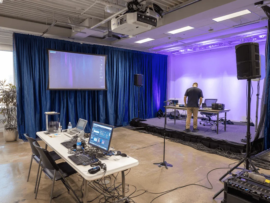 Indoor event setup with a small stage, a person working at a table with laptops, audio equipment, and a projector screen with blue curtains in the background.