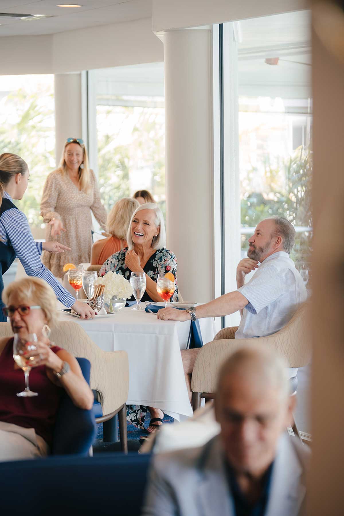 People dining and chatting at table in a bright restaurant with large windows and drinks on table.