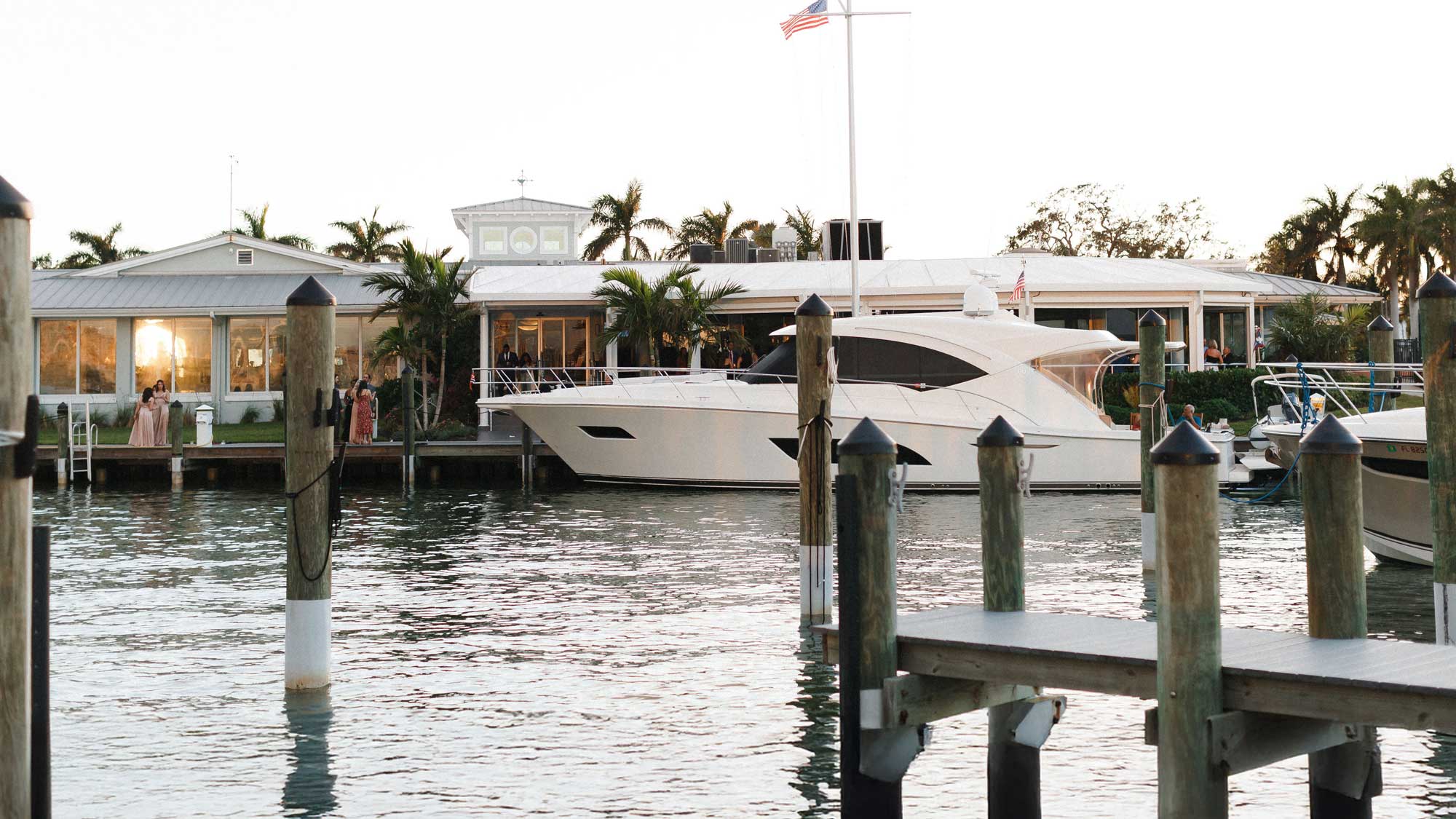 White yacht docked in calm harbor water near a building with palm trees and people.