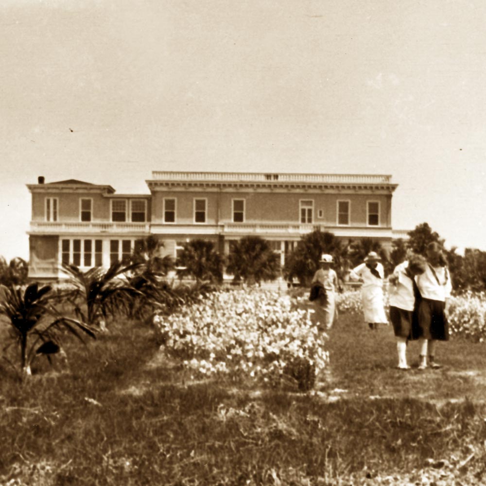 Sepia photo of four people walking on a grassy field with flowers in front of a large two-story building.