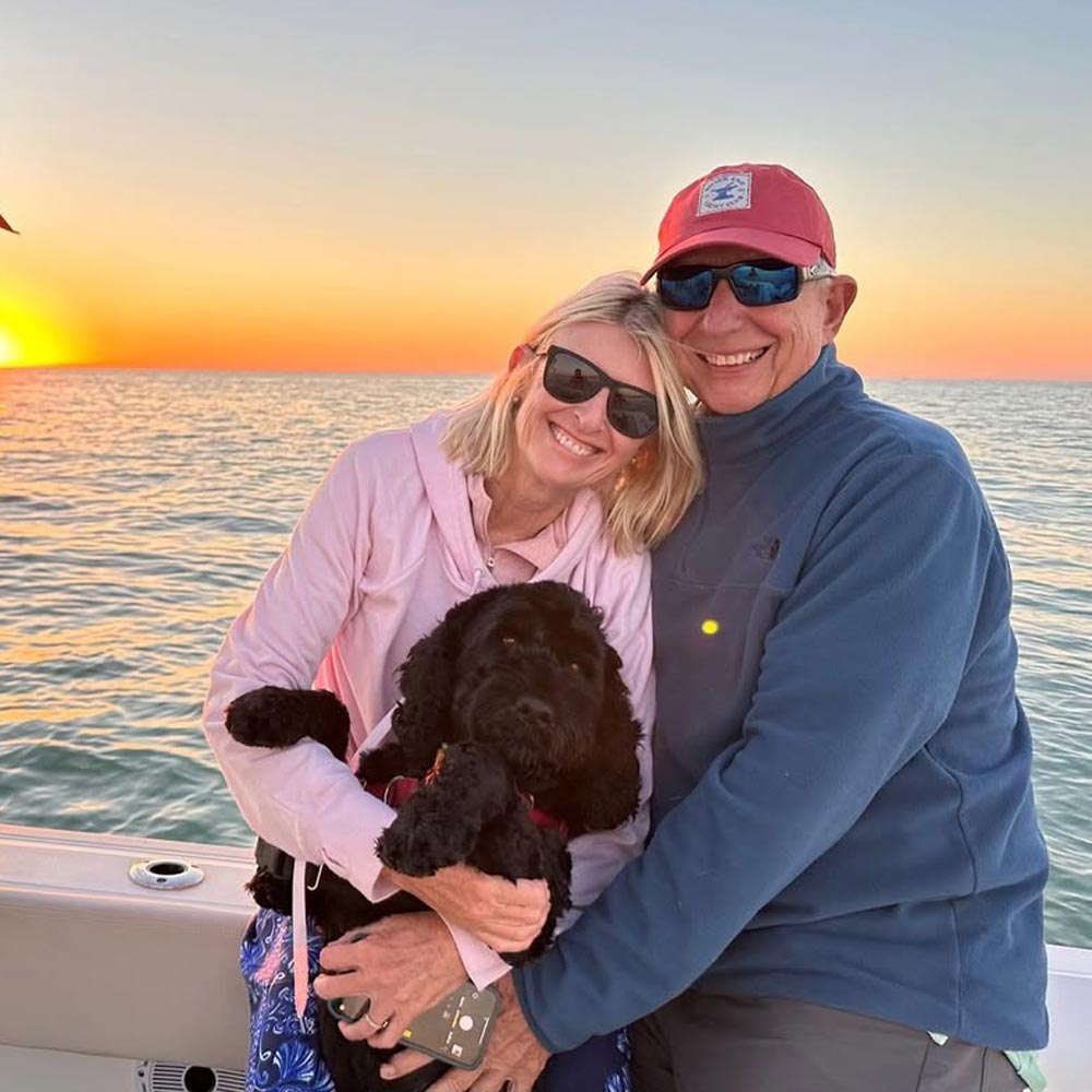 Smiling couple wearing sunglasses holding a black dog on a boat with a sunset over the ocean behind them.