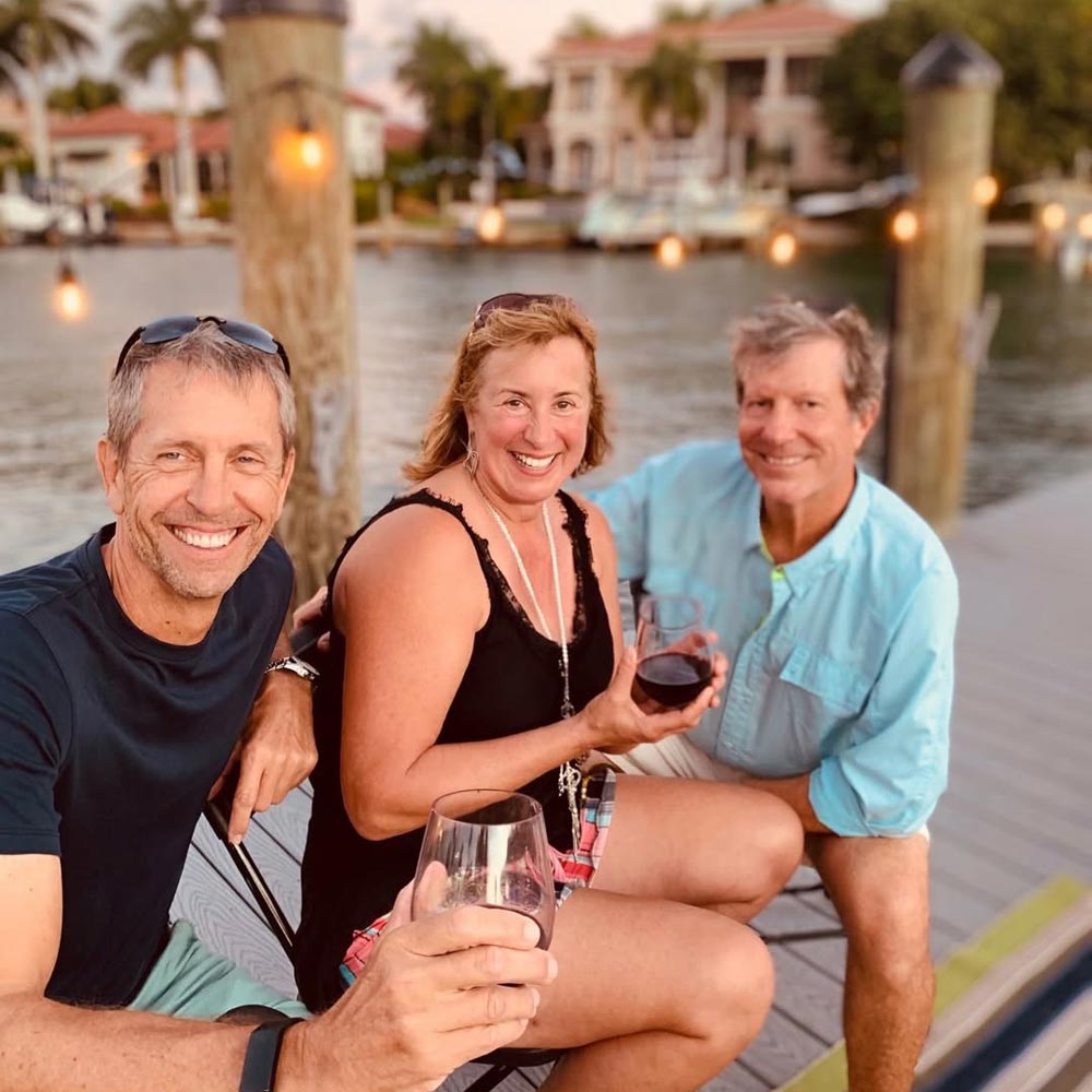 Three smiling adults sitting on a dock by the water holding wine glasses in a relaxed outdoor setting.