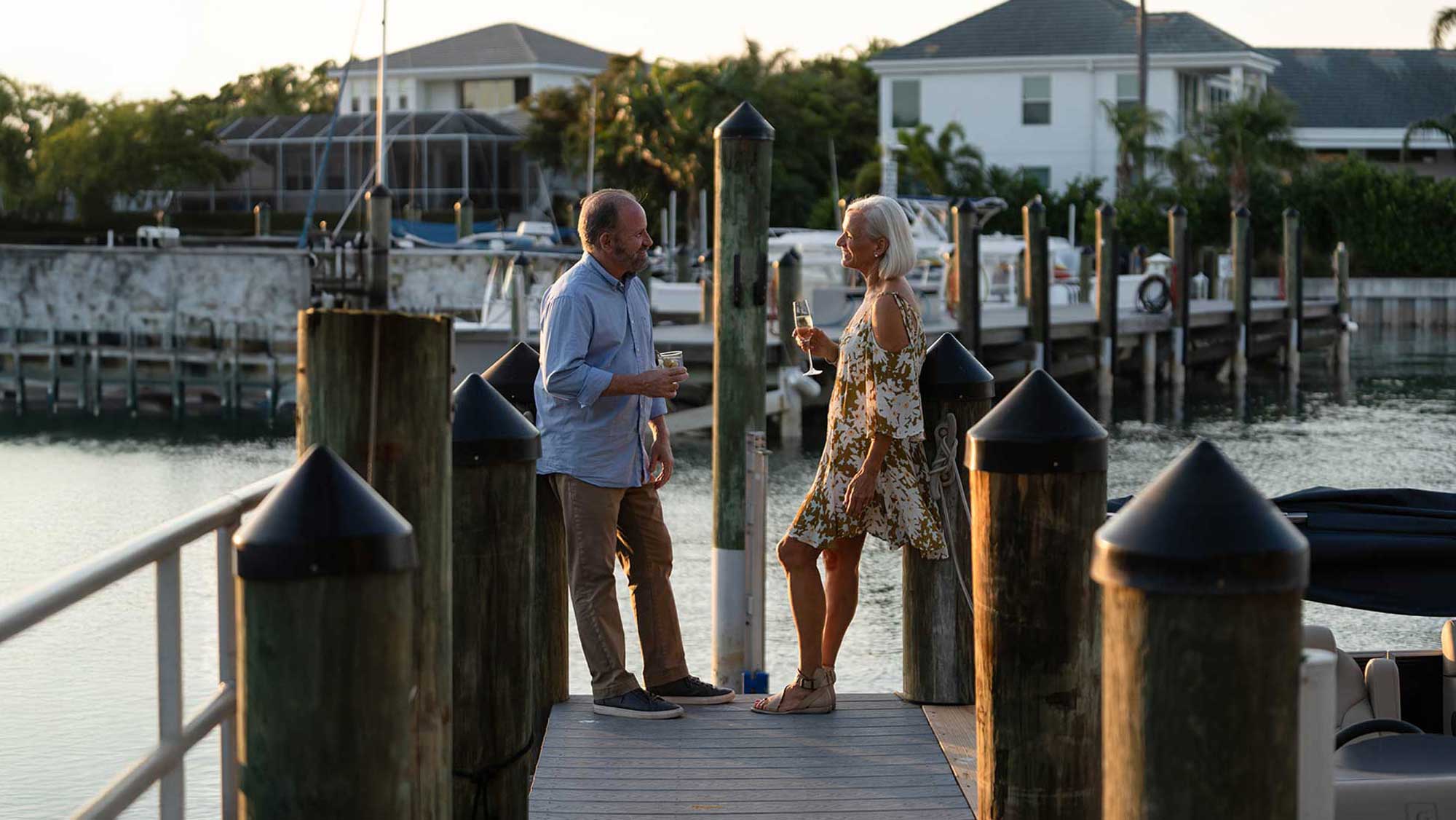 Older man and woman on a dock holding drinks and talking at sunset by waterfront homes.