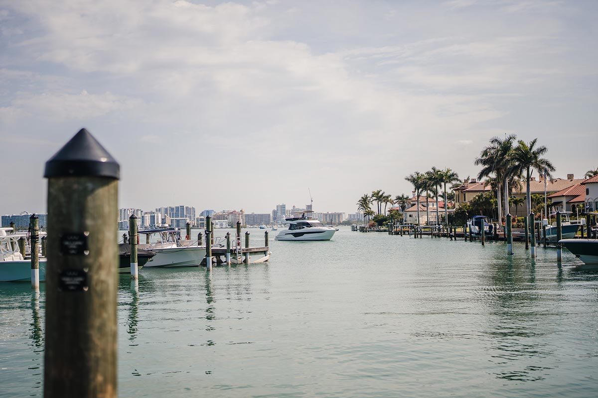 Waterway view of boats docked near palm trees and houses with a city skyline in the background.