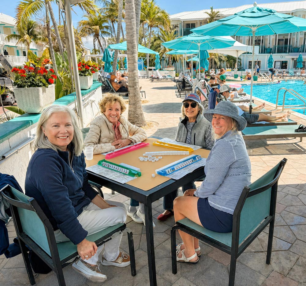 Four women sitting outdoors at a table playing Mahjong near a pool with palm trees and umbrellas.