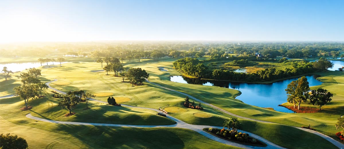 Aerial view of a golf course with green fairways, sand bunkers, trees, and water hazards under a clear sky.