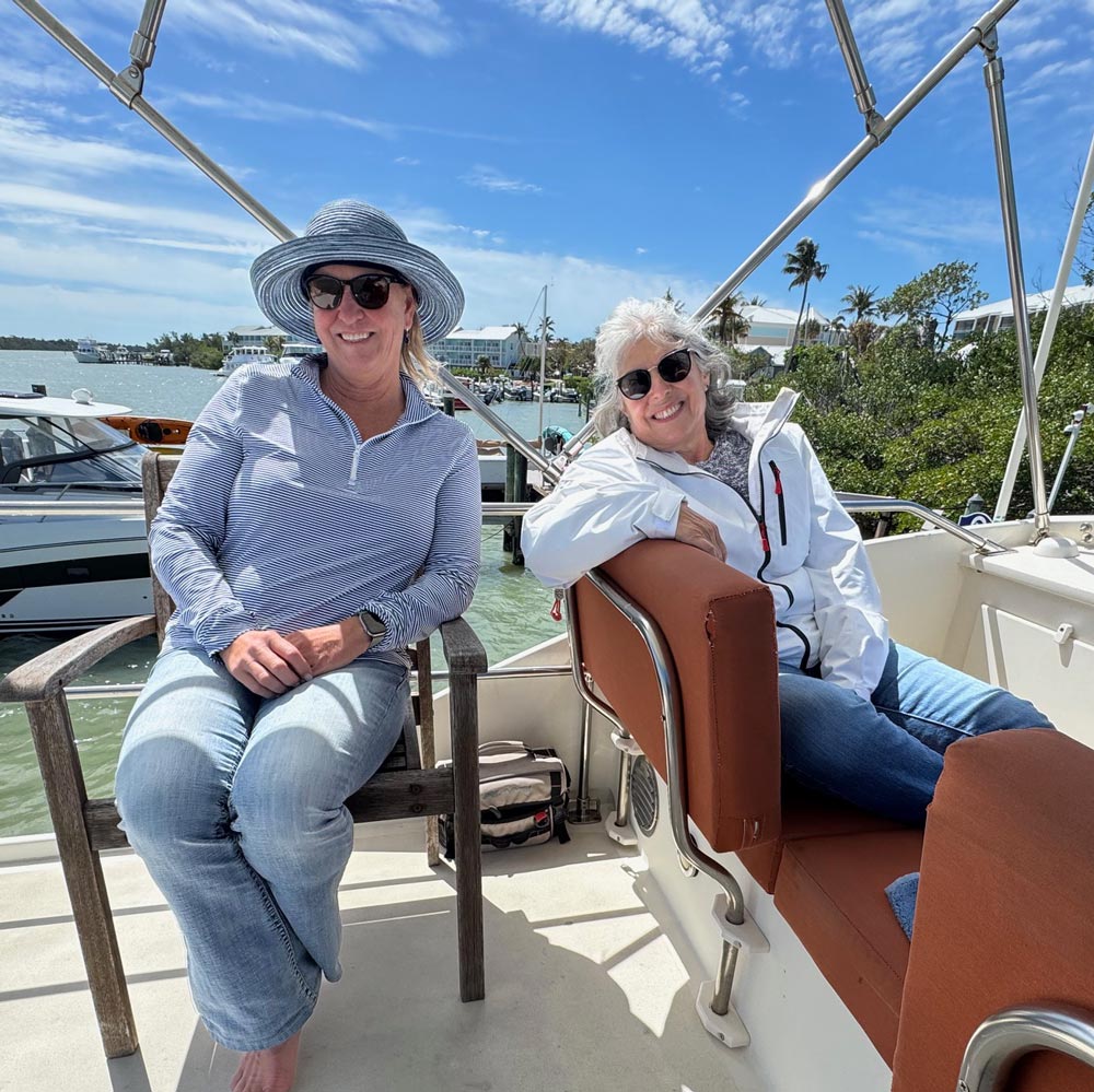 Two women wearing sunglasses, seated on a boat with water and buildings in the background under a blue sky.