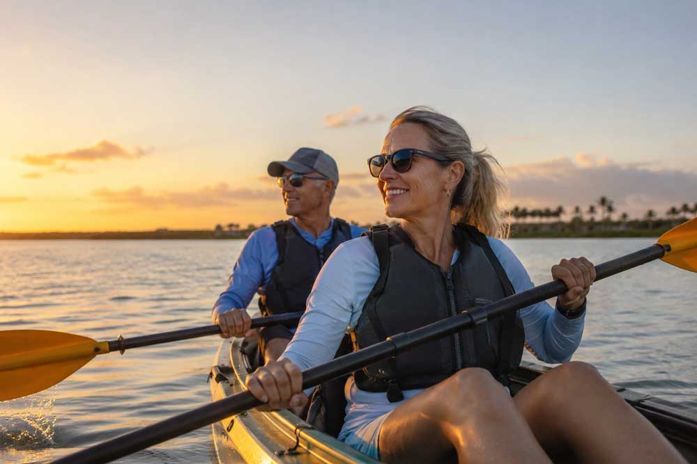 A man and woman wearing life vests kayaking together on calm water at sunset.