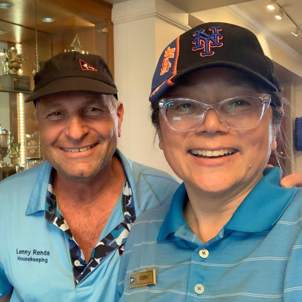 Two smiling people wearing blue shirts and hats standing indoors near a display case with trophies.