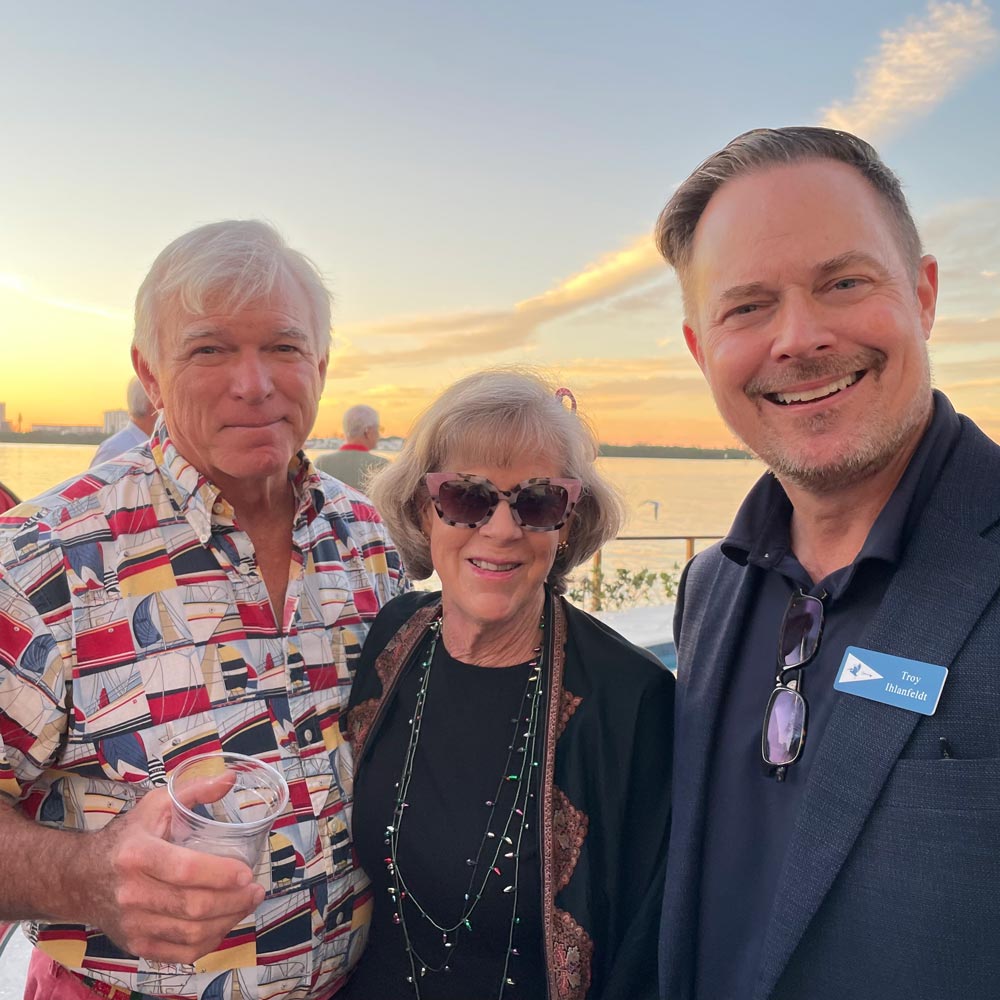 Three people smiling outdoors near water at sunset; man on right wears name tag 'Troy Ilhanfeldt'.