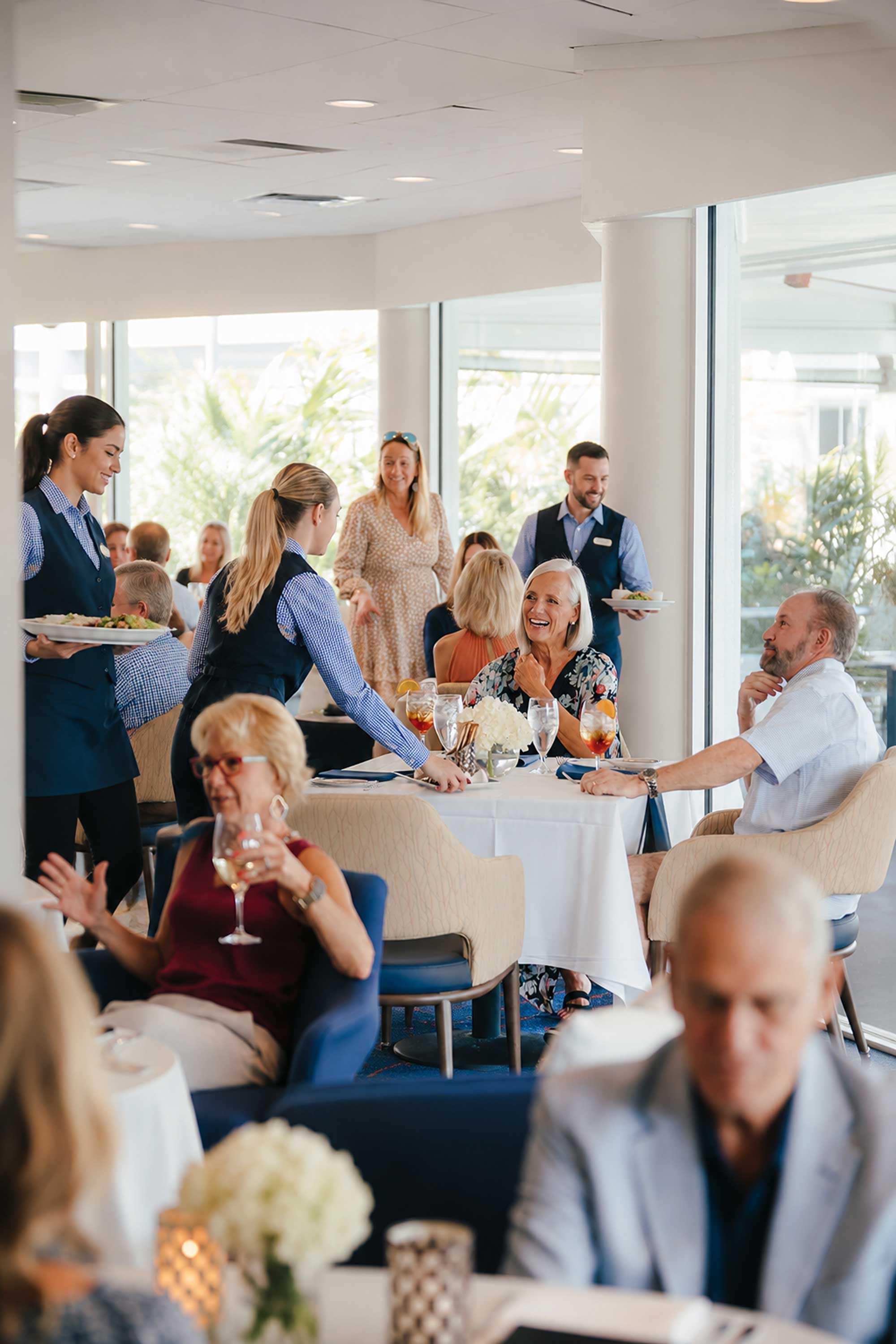 Waitstaff serving food in a bright, busy restaurant with seated diners enjoying meals and drinks.