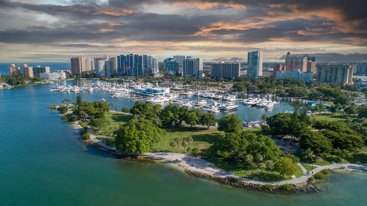 Aerial view of a marina with boats, waterfront park, and city skyline under a cloudy sky at sunset.