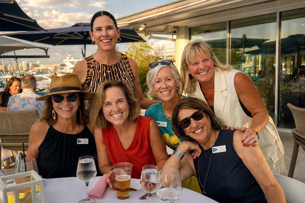 Six smiling women seated and standing around a table with drinks at an outdoor restaurant or event.