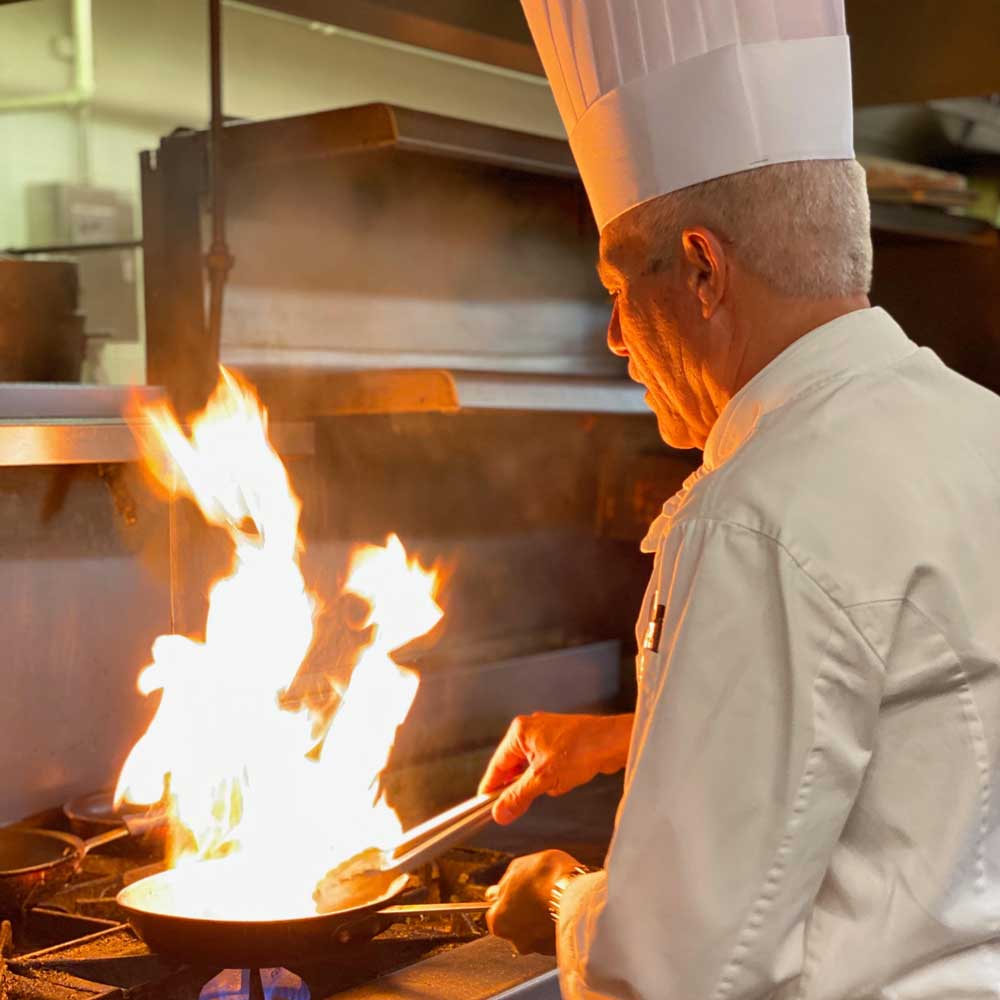 Chef in white uniform cooking with large flames on a stove in a professional kitchen.