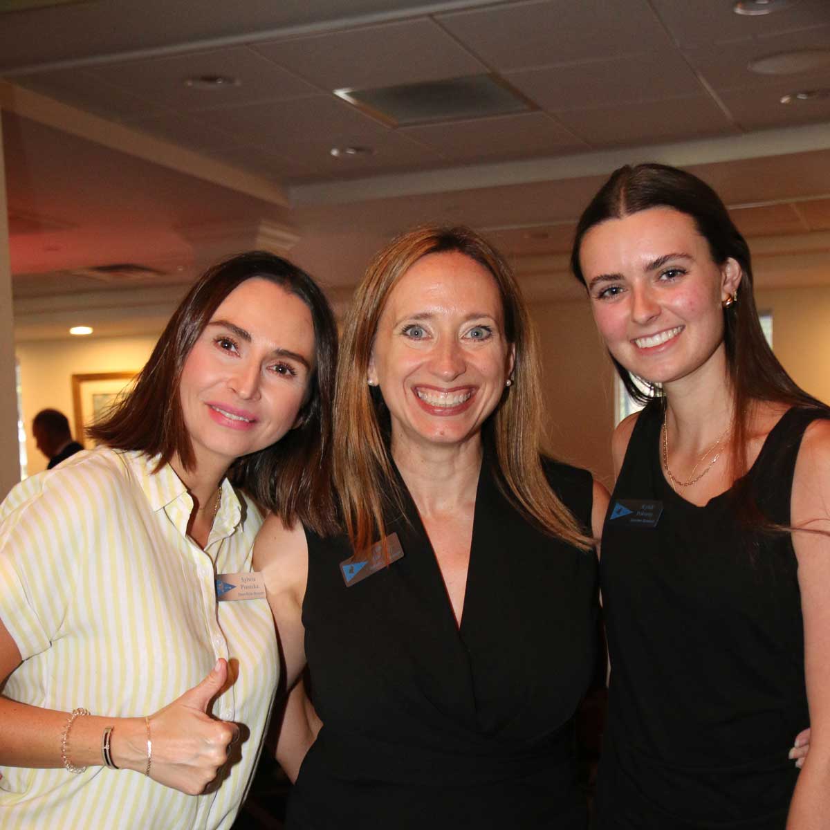 Three women smiling indoors, one giving a thumbs-up, all wearing name tags and casual attire.