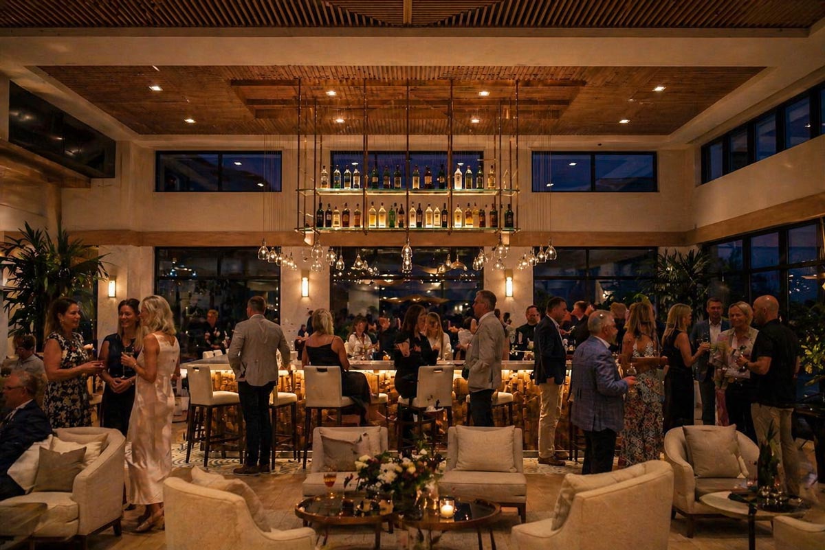 People socializing and drinking in a warmly lit, modern bar with cream furniture and hanging lights.