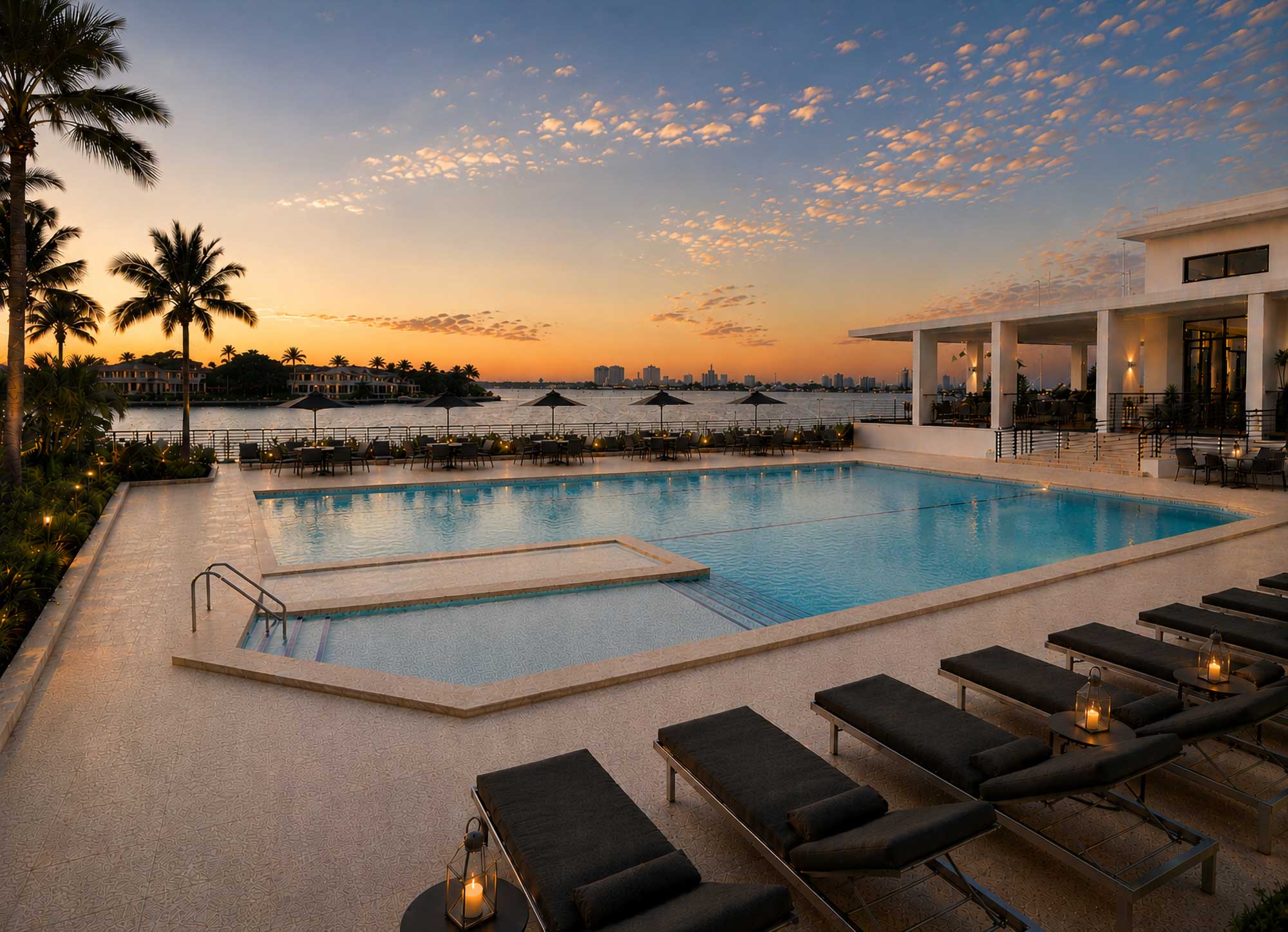 Outdoor pool with lounge chairs and umbrellas at sunset overlooking water and city skyline.