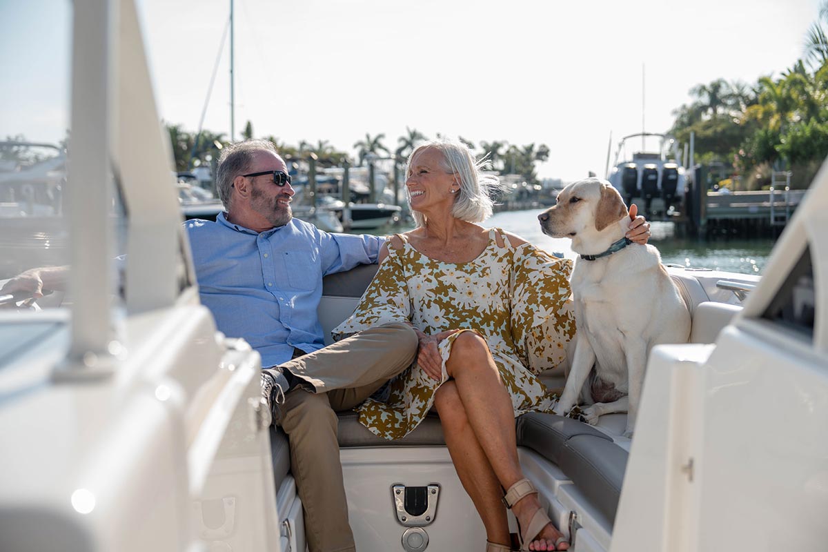 An older couple sitting on a boat with their dog in a marina on a sunny day.