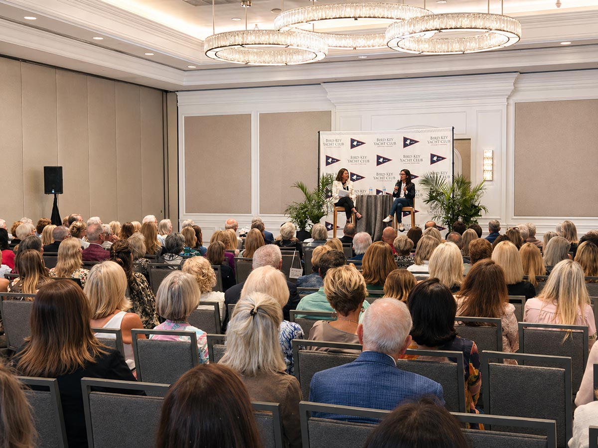 Audience seated in a conference room watching two women speaking on stage at Bird Key Yacht Club event.