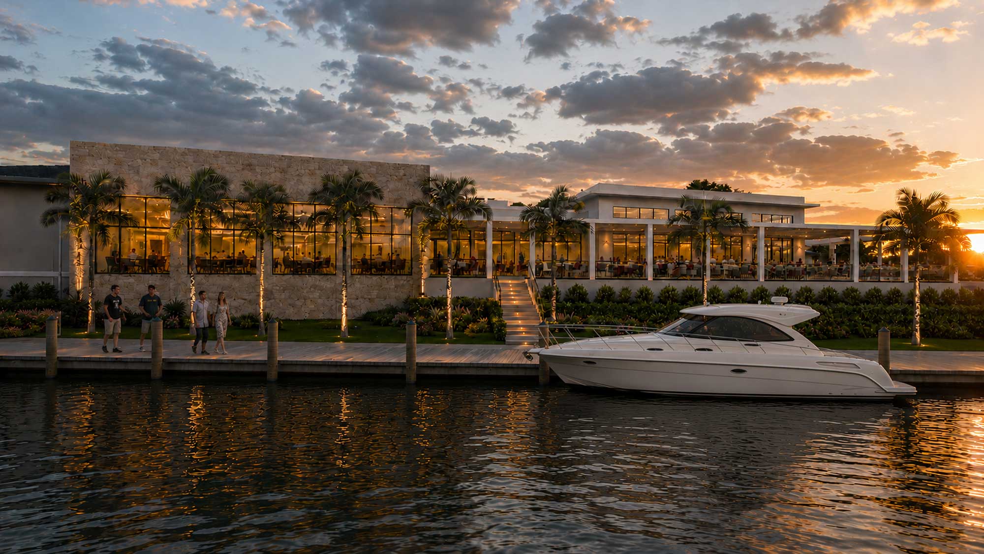 Modern waterfront building with illuminated palm trees, people walking, and a docked white yacht at sunset.