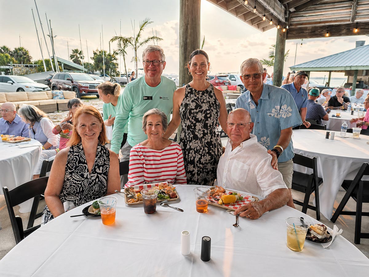 Group of six adults smiling around a table with food and drinks at an outdoor event.