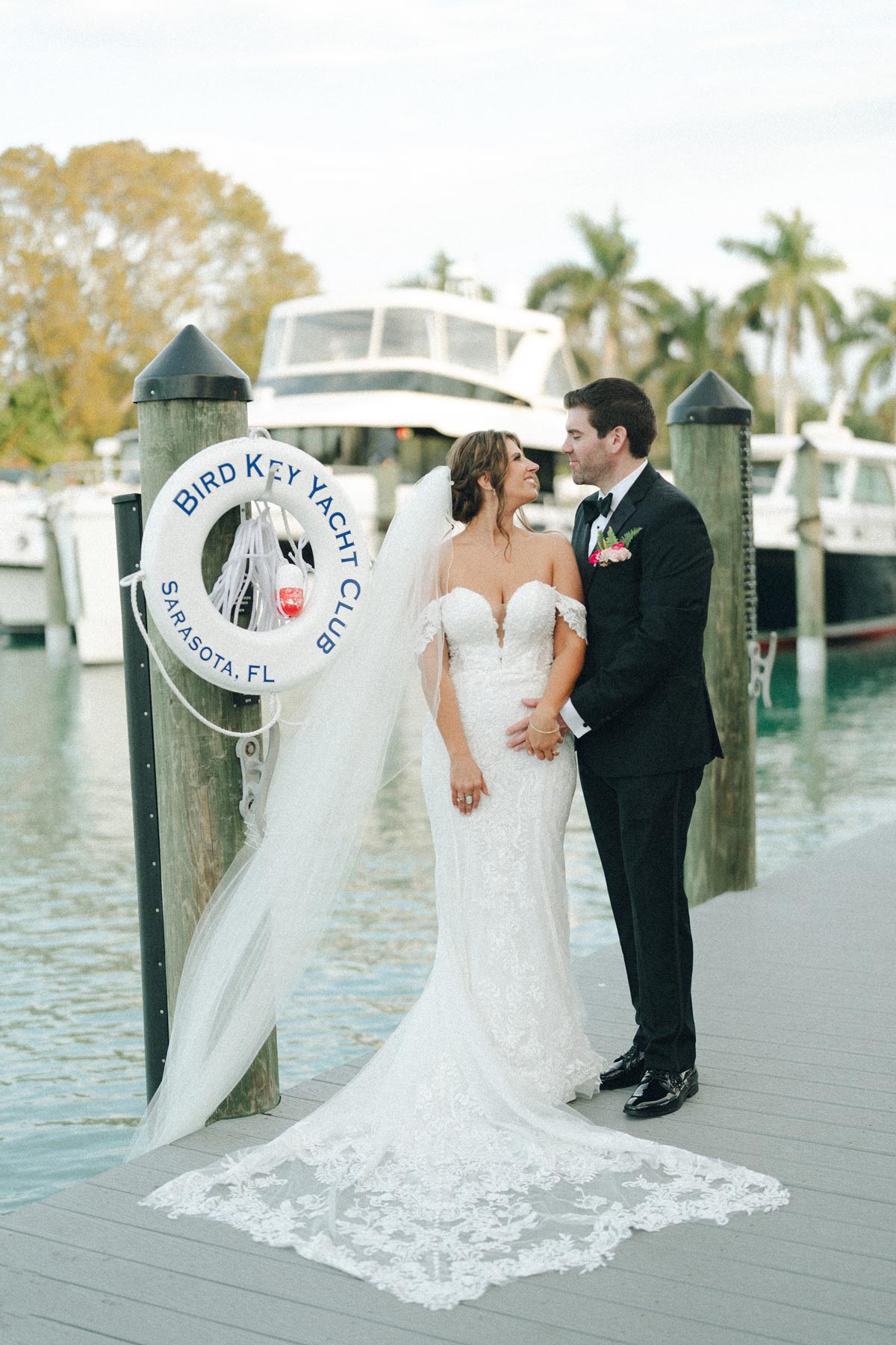 Bride and groom smiling on dock at Bird Key Yacht Club with boats and palm trees in background.