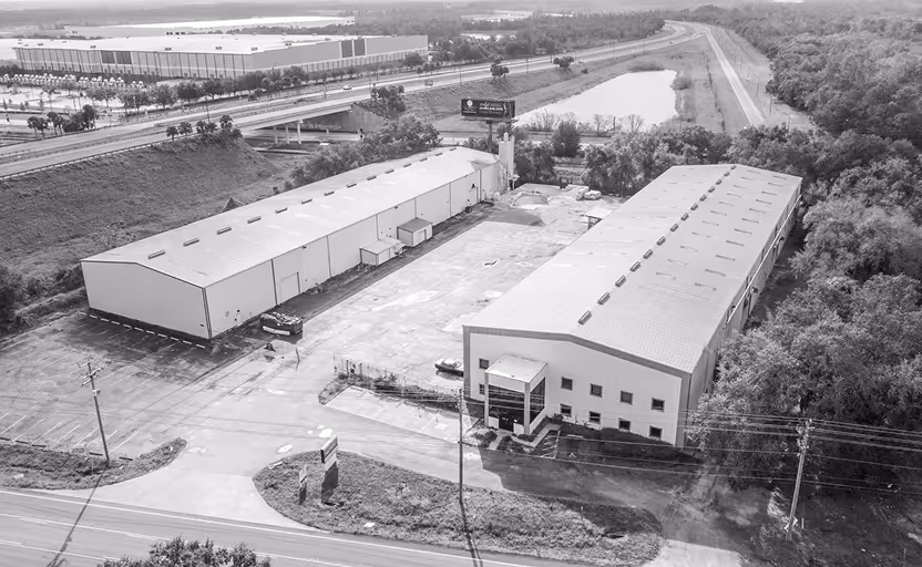 Aerial view of two large industrial warehouse buildings separated by a paved lot near highways and surrounded by trees.