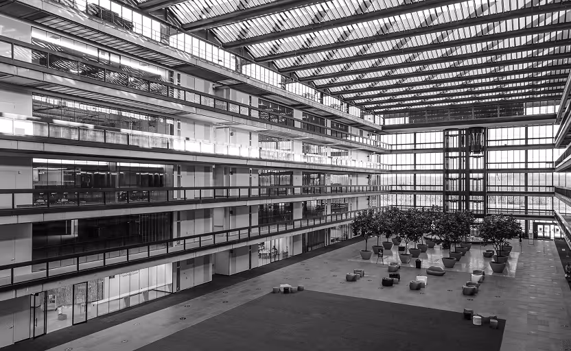Spacious modern atrium with glass ceiling, multiple floors of offices, potted plants, and scattered seating areas.