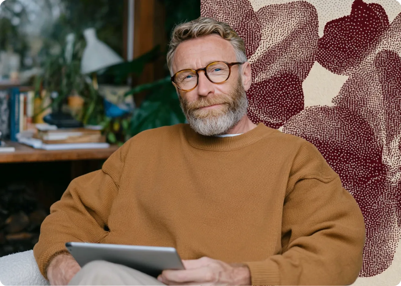 Homme mûr avec lunettes et pull marron tenant une tablette, assis dans un fauteuil blanc.