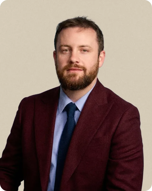 Homme avec barbe portant un costume bordeaux, chemise bleue et cravate bleue devant un fond beige.