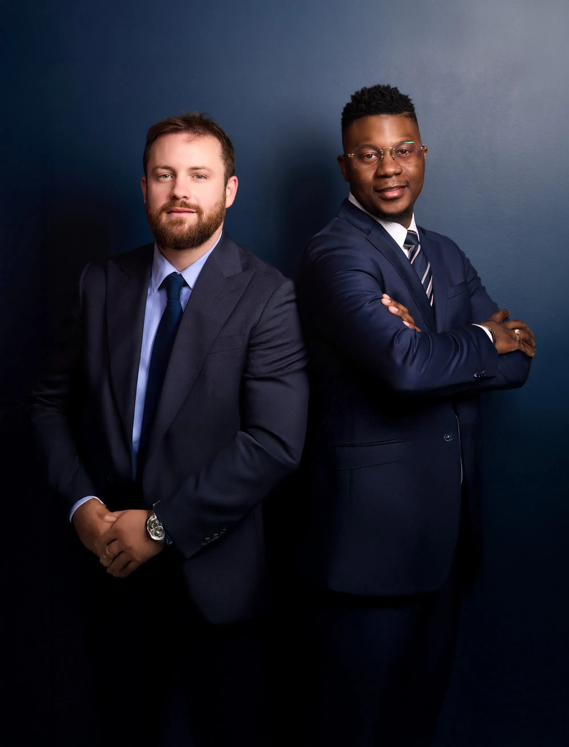Deux hommes en costume professionnel posent debout côte à côte sur fond bleu foncé.