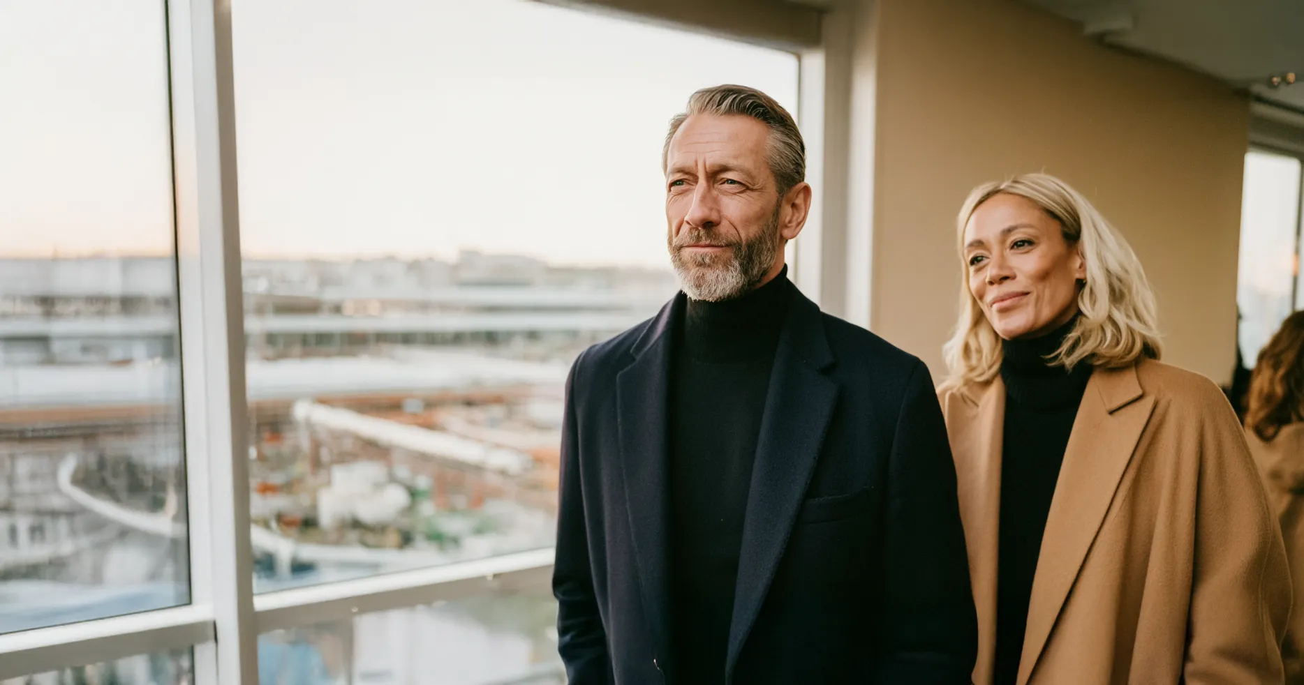 Un homme avec une barbe grise en manteau noir et une femme blonde en manteau camel regardant par une fenêtre.