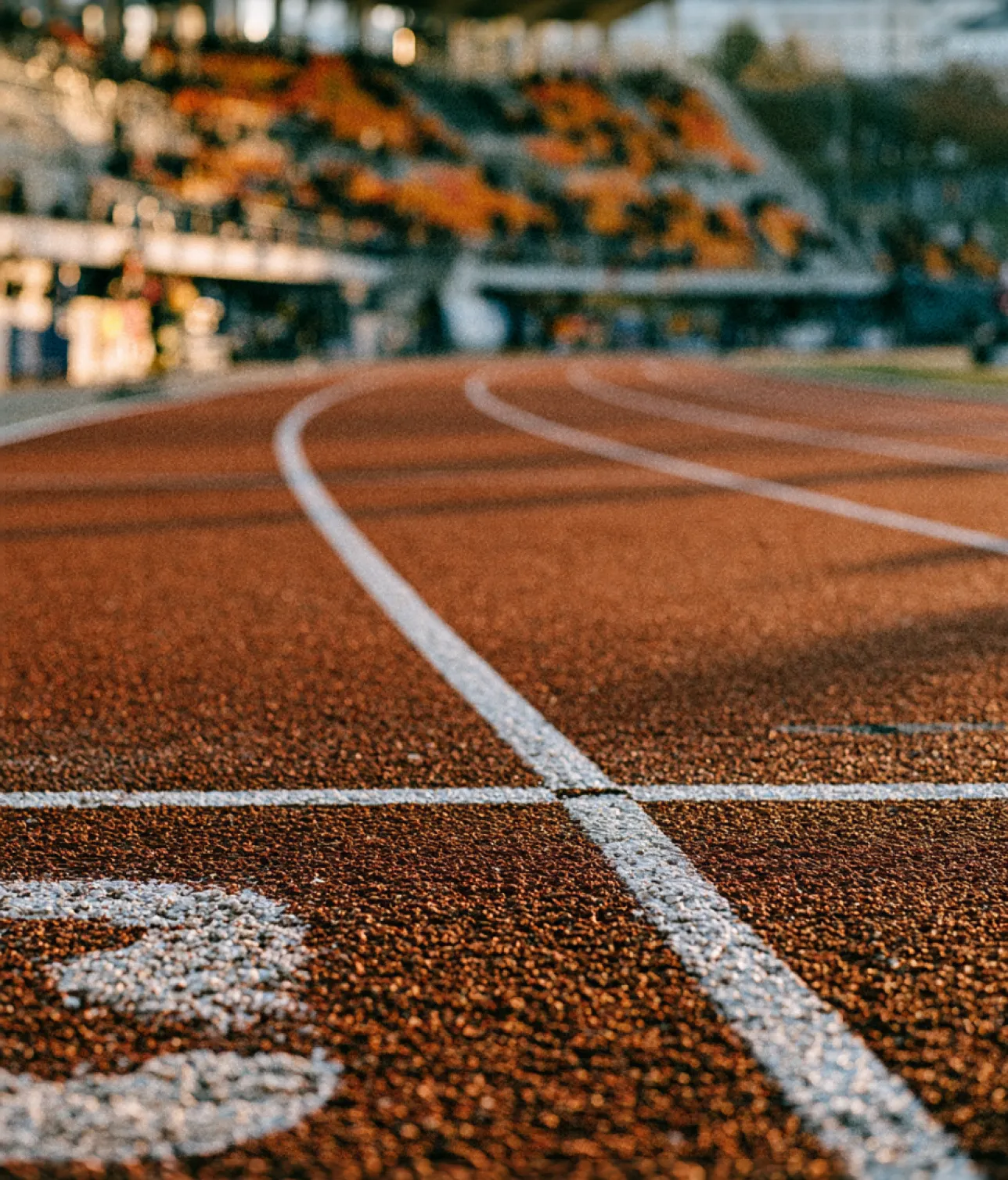 Piste d'athlétisme rouge vue de près avec lignes blanches courbées et gradins flous en arrière-plan.