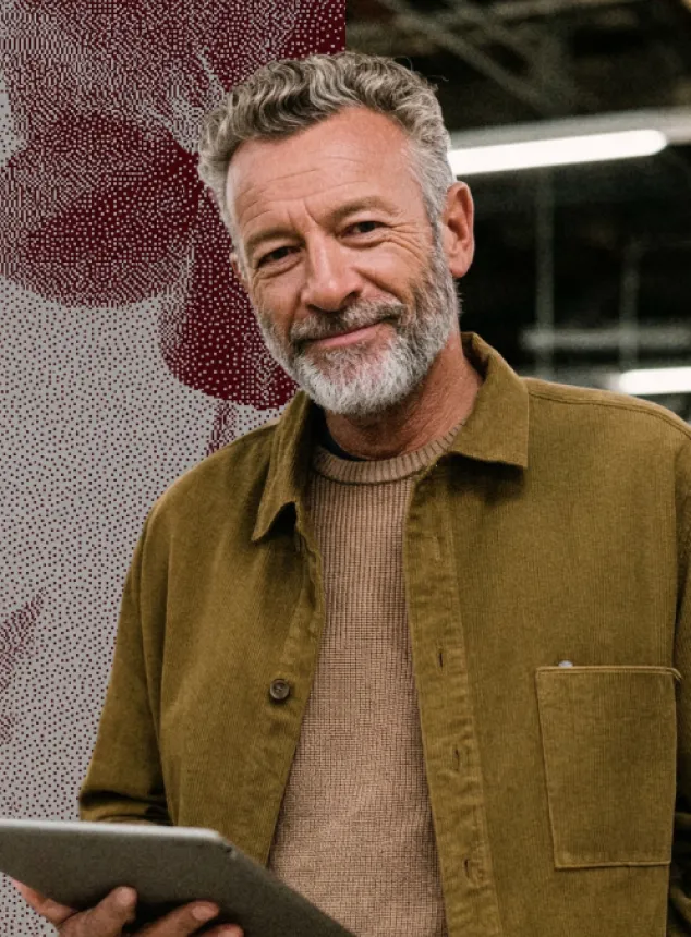 Homme souriant avec barbe grise, portant une veste marron et tenant une tablette.