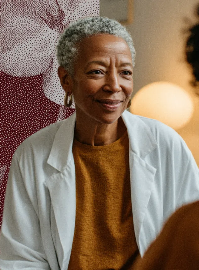 Une femme âgée souriante avec des cheveux gris courts porte un pull moutarde et une blouse blanche.