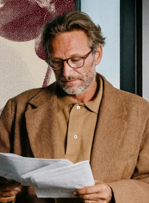 Homme portant des lunettes et un manteau marron lisant un journal plié.