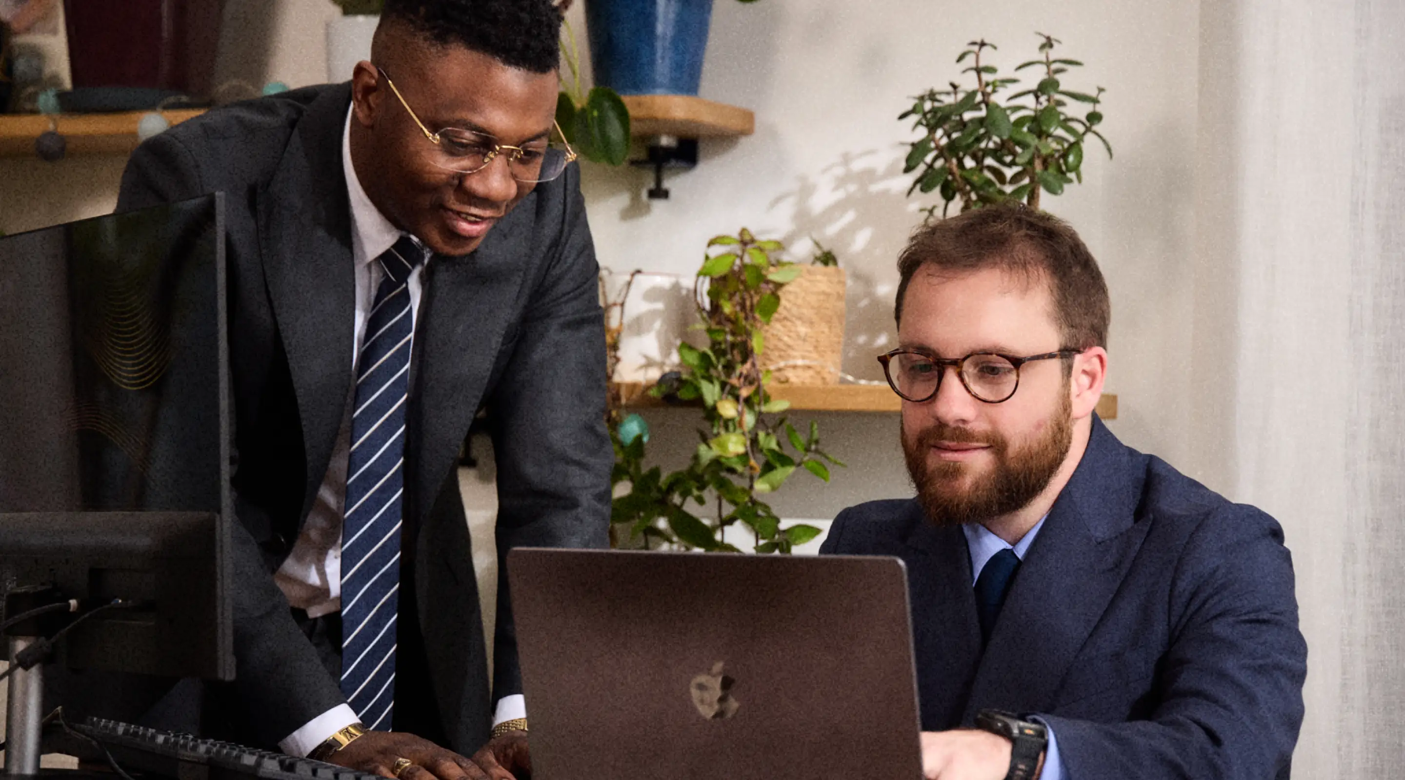 Deux hommes en costume discutent devant un ordinateur portable dans un bureau avec des plantes.