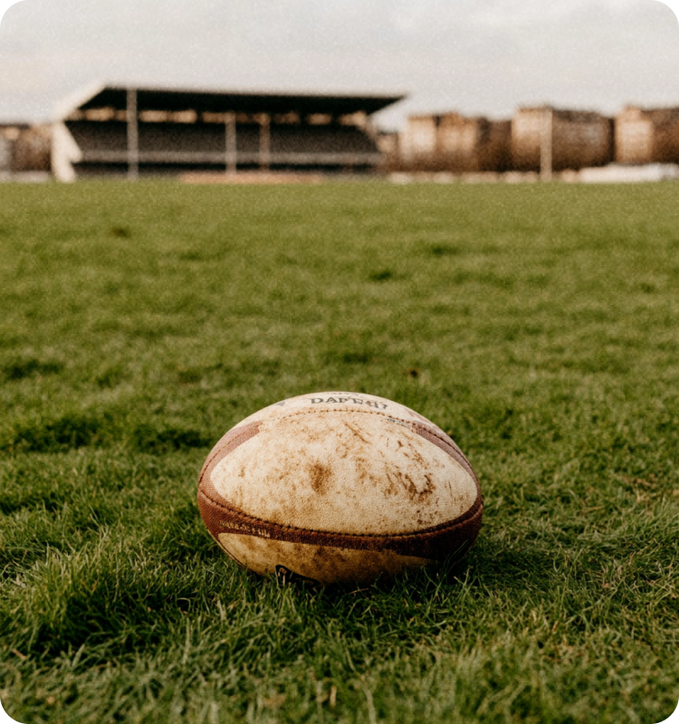 Un ballon de rugby usé posé sur un terrain d'herbe avec des tribunes en arrière-plan.