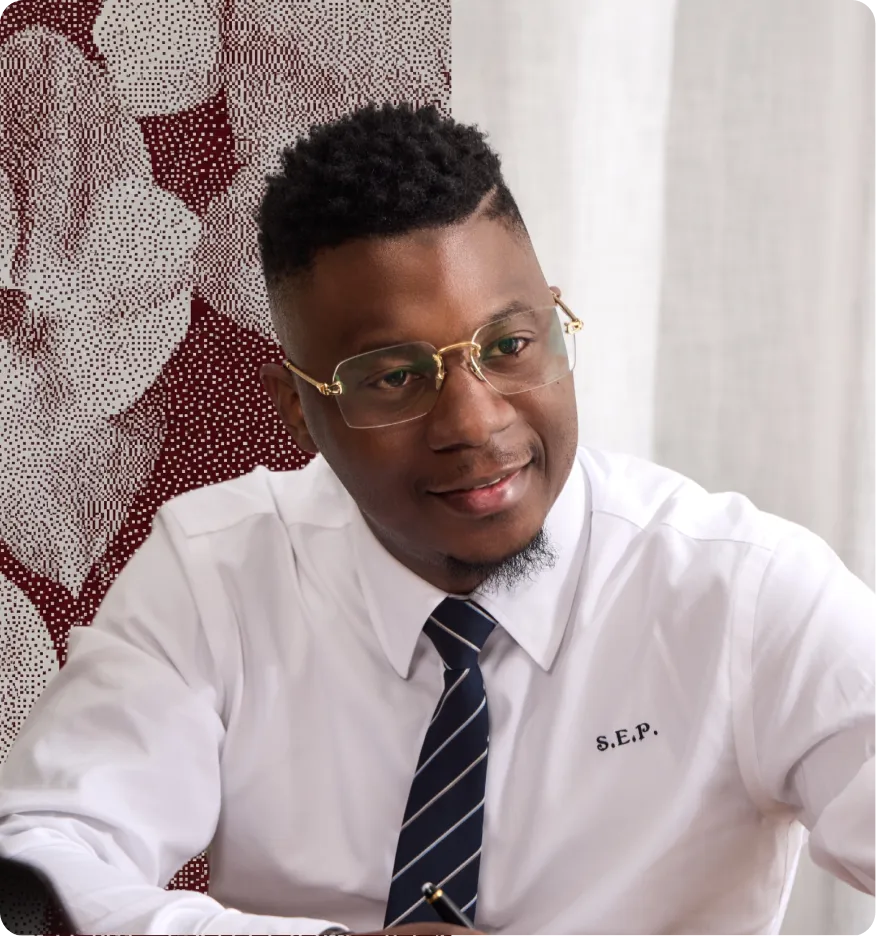 Homme noir souriant, chemise blanche avec S.E.P., cravate, et lunettes assis devant une fenêtre.