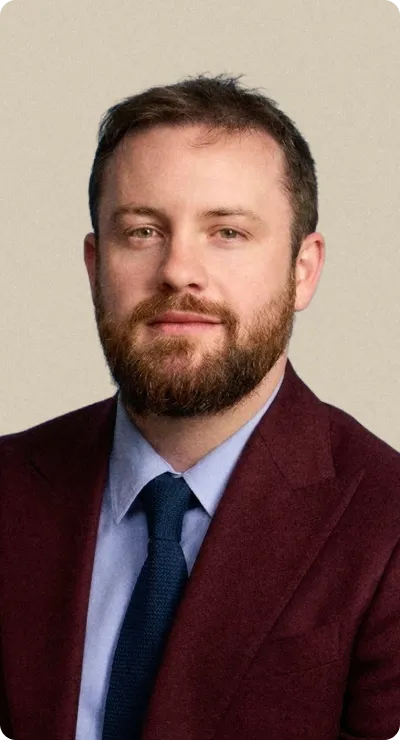 Portrait d'un homme avec barbe portant un costume bordeaux, chemise bleue et cravate noire.