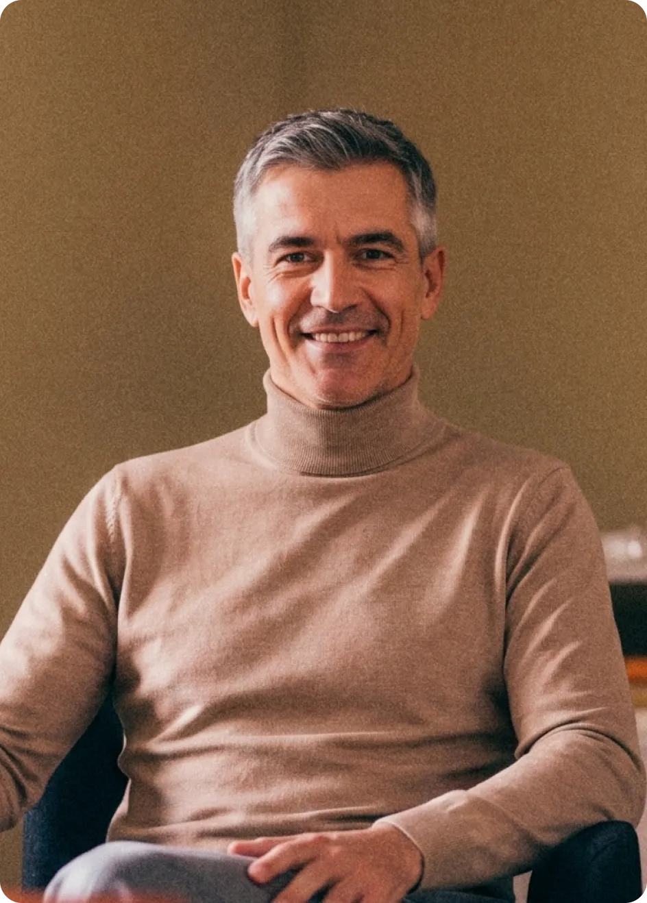 Homme souriant aux cheveux grisonnants portant un col roulé beige assis confortablement.