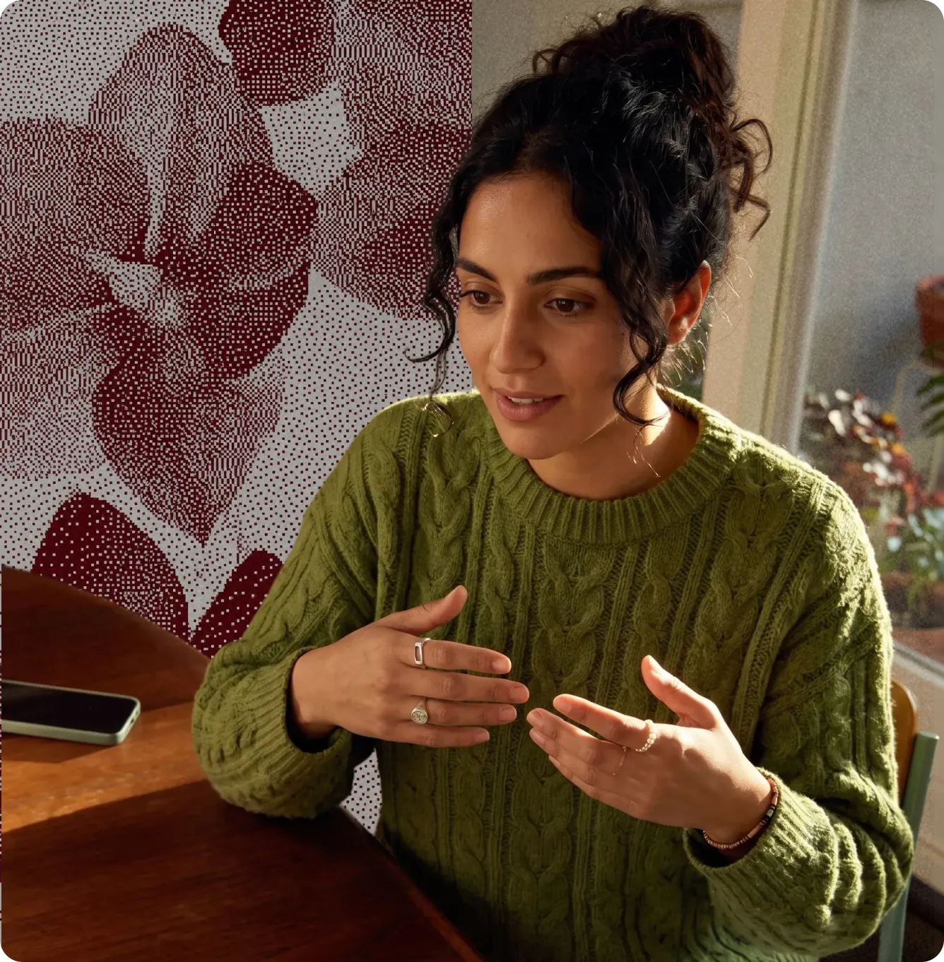 Femme en pull vert parlant avec ses mains posée à une table en bois avec un téléphone à côté.