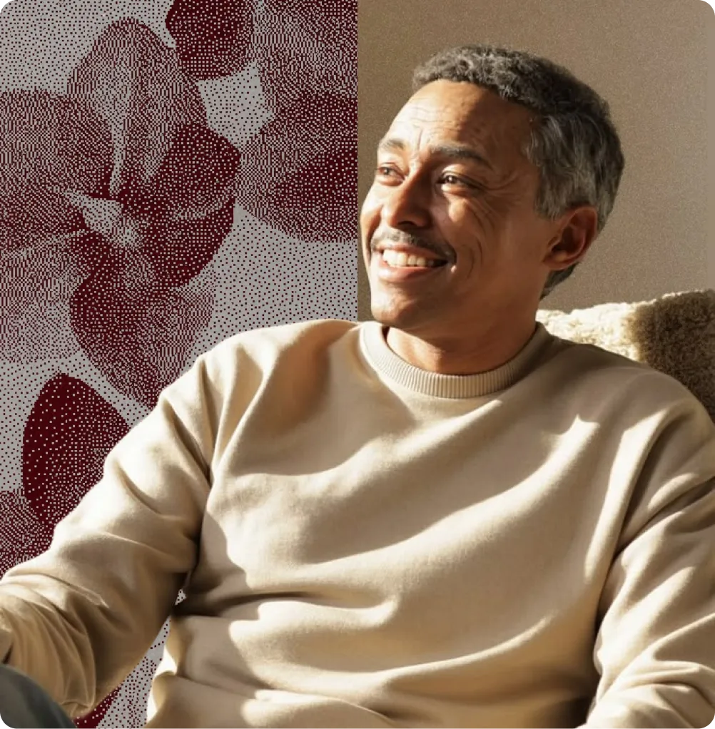 Homme souriant assis près d'un mur beige avec un motif floral rouge en arrière-plan.